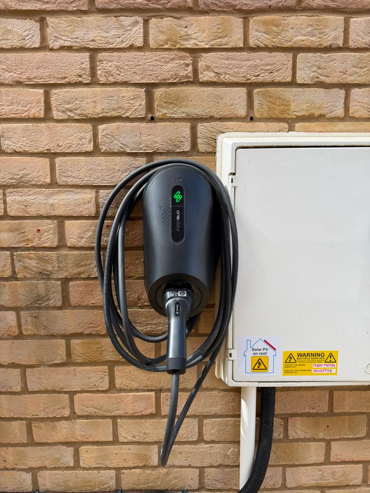 A myenergi Zappi Glo EV charger mounted on a brick wall next to an isolator
